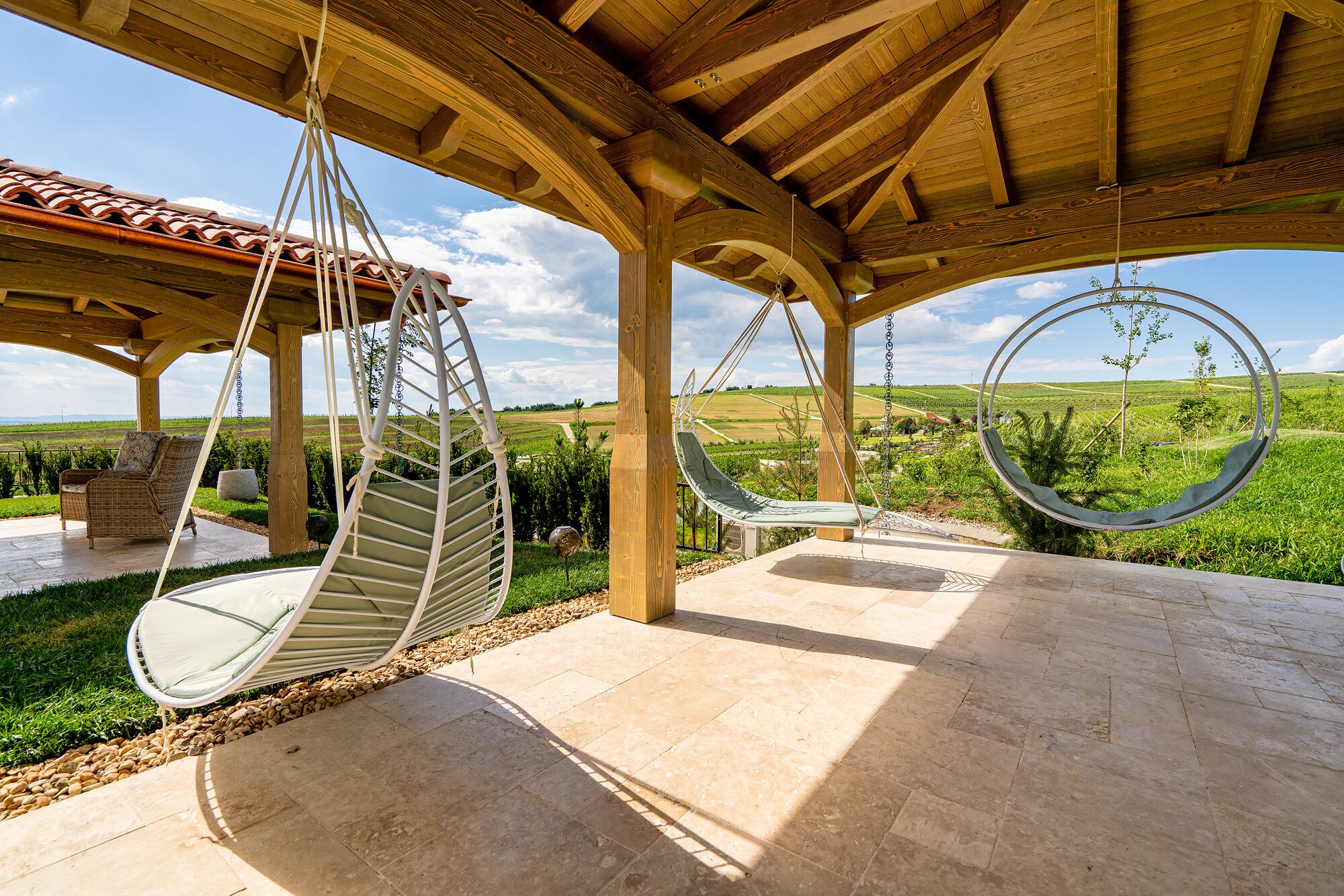 A covered outdoor area with two swing chairs, a wicker chair, and a view of a vineyard.