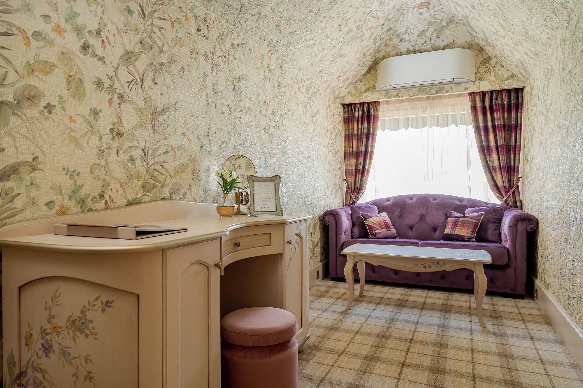 A room with floral wallpaper, a purple sofa, a small table, and a desk with drawers.