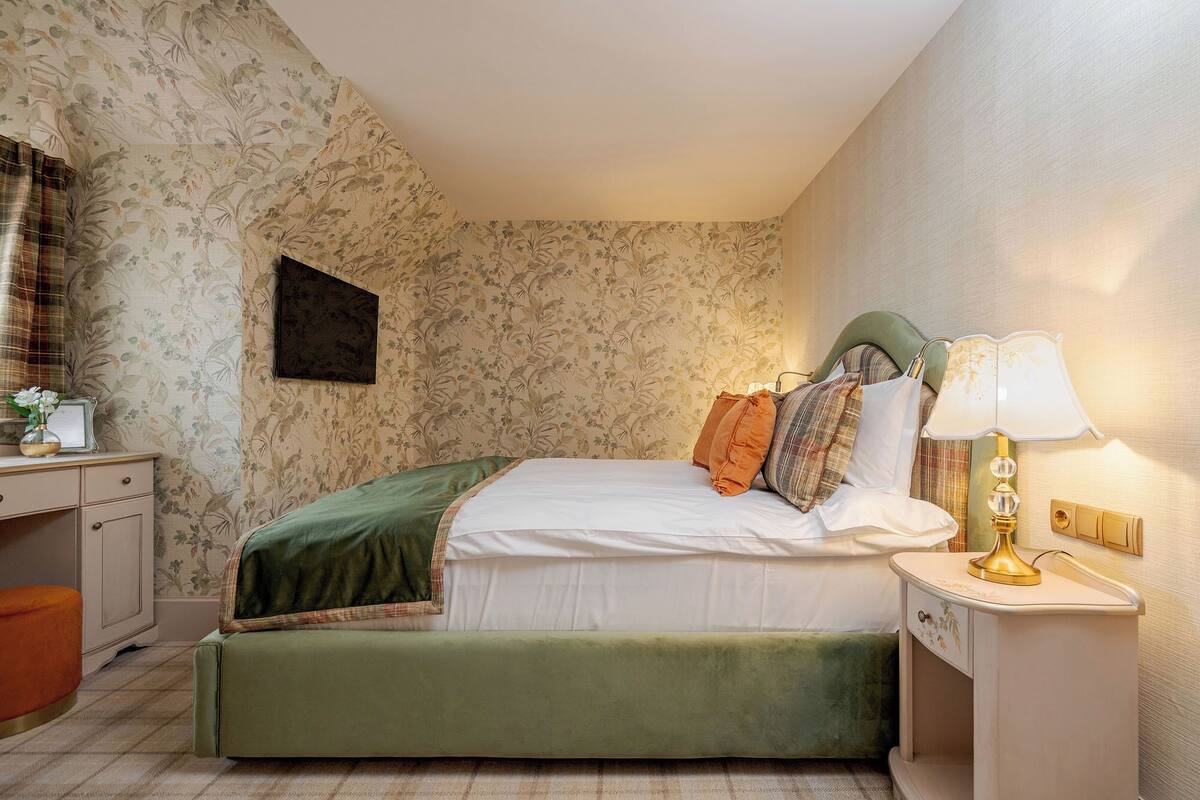 A bedroom with floral wallpaper, a green upholstered bed, a bedside table with a lamp, and a small desk with a vase of flowers.