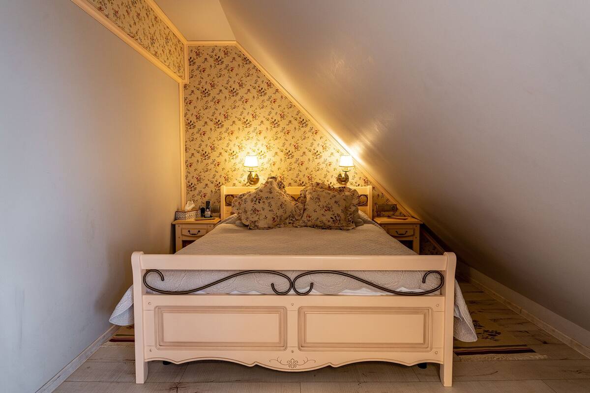 A bedroom with a sloped ceiling, floral wallpaper, a bed with a decorative headboard, two bedside tables with lamps, and a nightstand with a lamp.