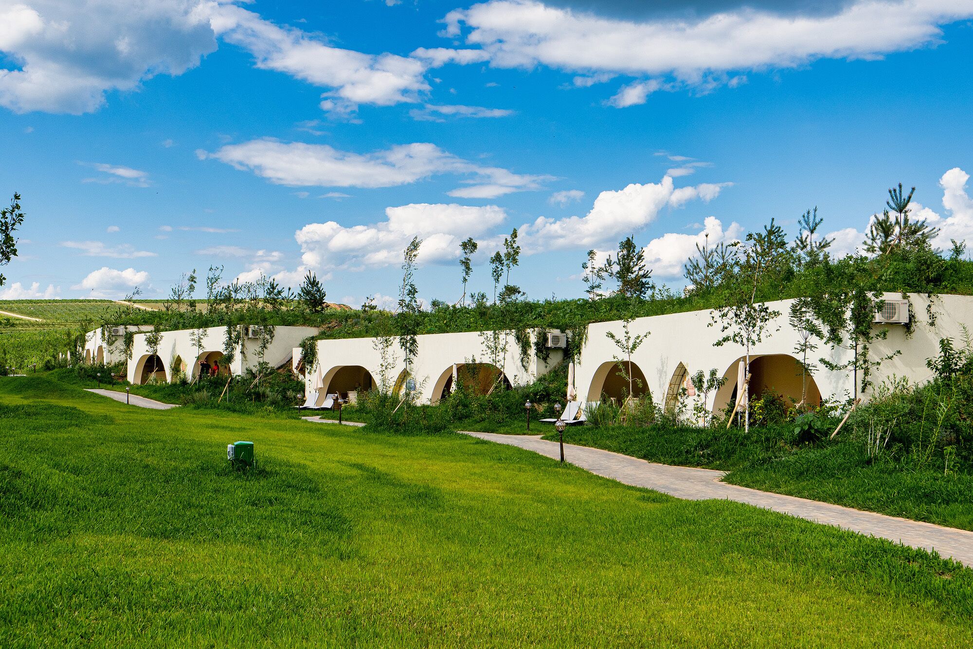 Une rangée de structures blanches en forme d’arc, reliées par un chemin, entourées de verdure et surplombées par un ciel dégagé.