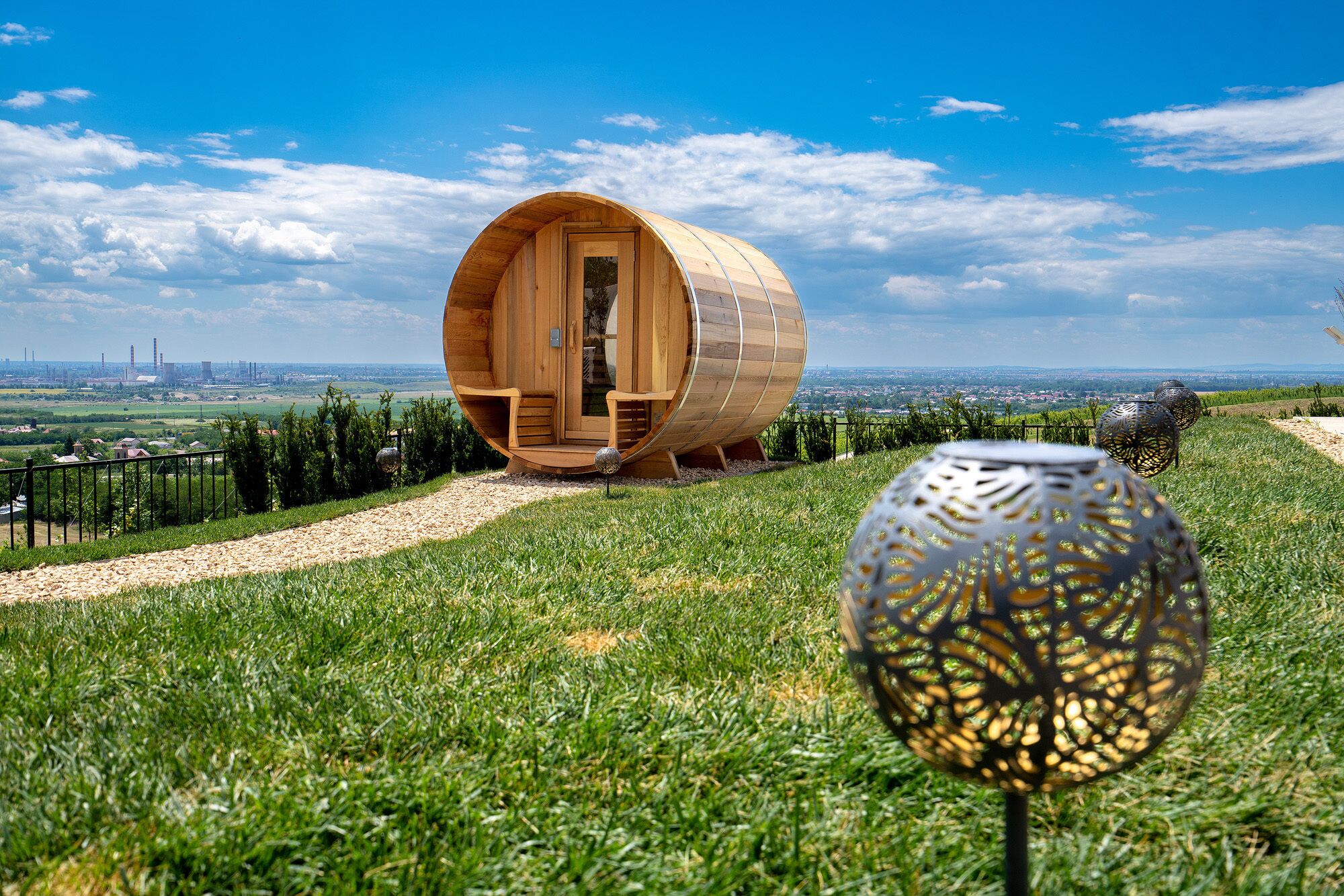 A wooden barrel-shaped structure with a door and seating area, surrounded by a grassy landscape and a clear sky.