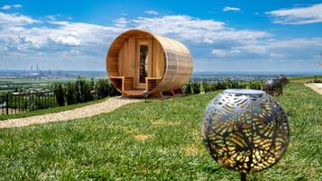 A wooden barrel-shaped structure with a door and seating area, surrounded by a grassy landscape and a clear sky.
