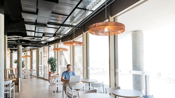 Un café moderno con mesas redondas y lámparas colgantes, con amplias ventanas y un espacio exterior visible.