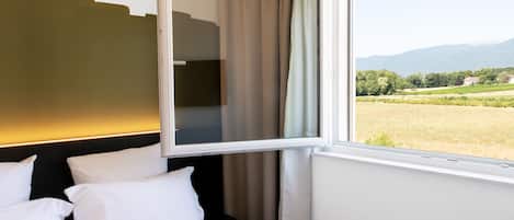 A hotel room with a bed, white pillows, a window with a view of a field and mountains.