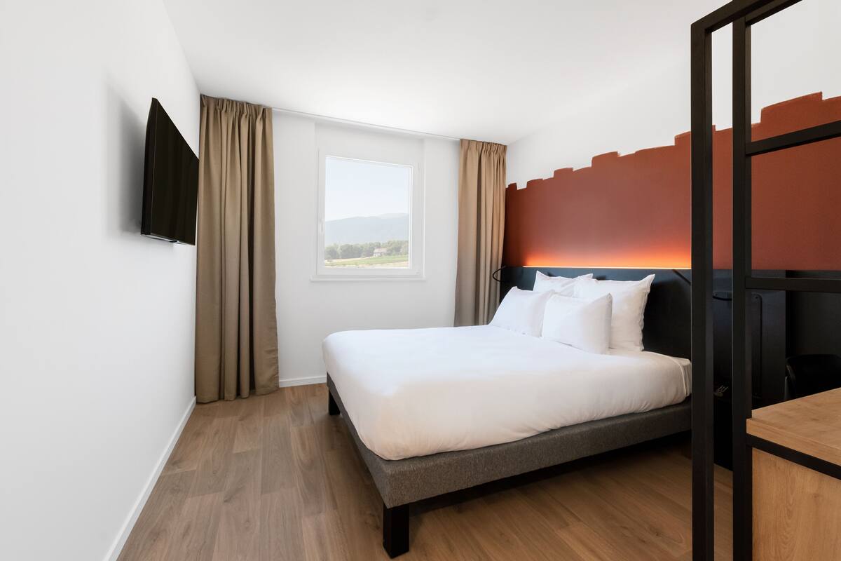 A modern hotel room with a bed, a TV mounted on the wall, a window with curtains, and a view of the outdoors.