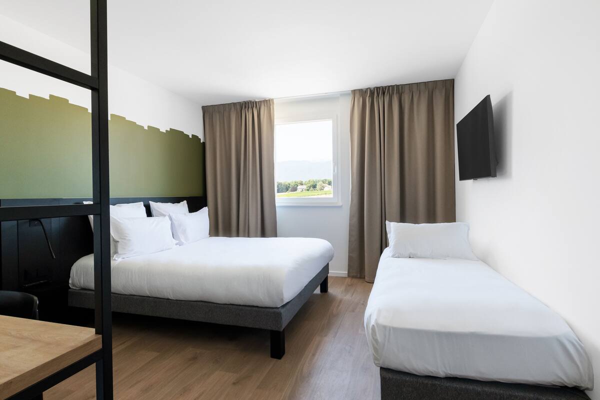 A hotel room with two single beds, a wooden desk, a TV mounted on the wall, and a window with curtains.