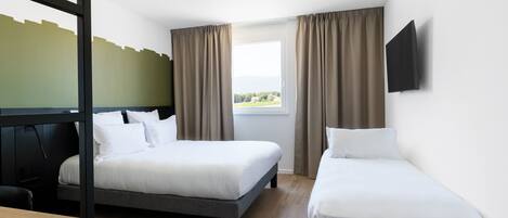 Habitación de hotel con dos camas individuales, un escritorio de madera, un televisor montado en la pared y una ventana con cortinas.