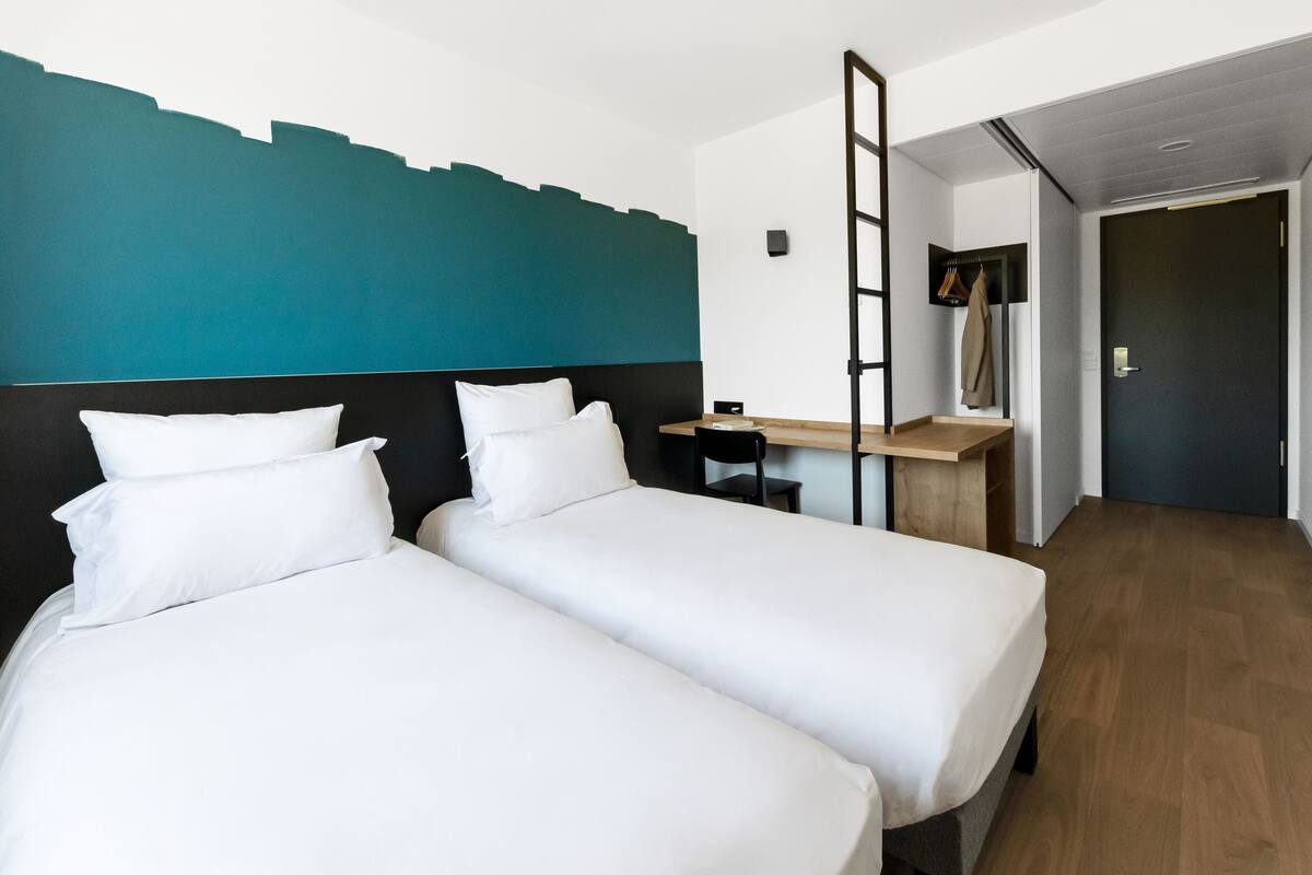 A hotel room with two single beds, a desk, a chair, and a TV mounted on the wall.