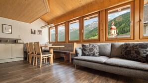 Un salon chaleureux avec un plafond en bois, un canapé agrémenté de coussins à motifs floraux, un coin repas avec des chaises et de grandes fenêtres offrant une vue sur les montagnes.