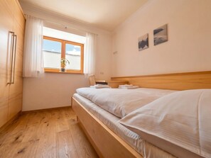 A bedroom with a wooden bed, a window with curtains, a plant, and two wall-mounted pictures. - In the Center of Neustift - TWO Bedroom Apartment - sleeps 6 (Neustift im Stubaital)