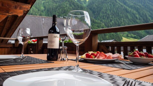 Une table dressée pour le dîner, avec une bouteille de vin, des verres et une assiette de nourriture, donnant sur un paysage montagneux.