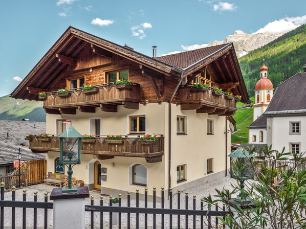 A traditional chalet with wooden balconies, a bell tower, and a mountain backdrop.
