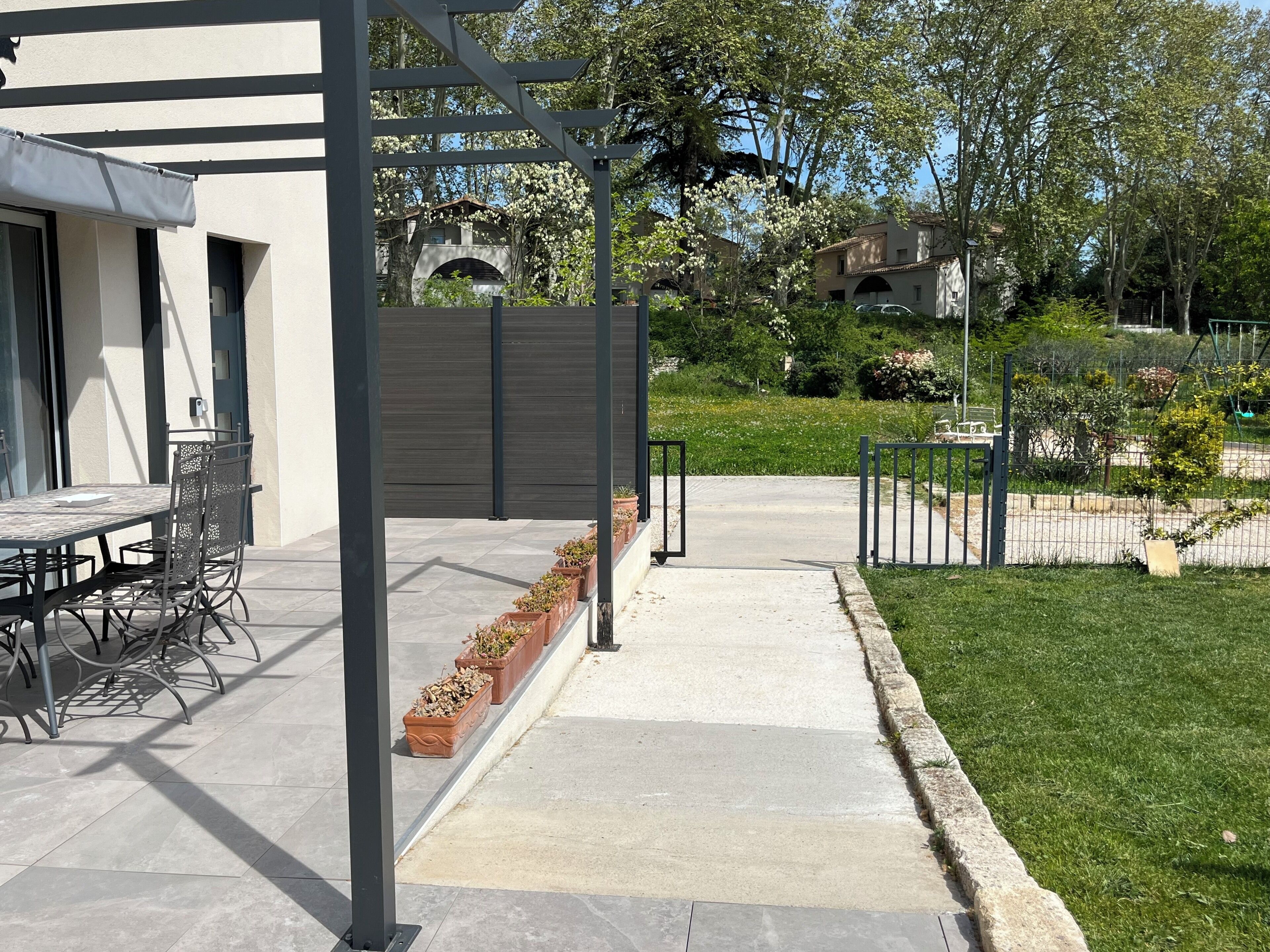 En uteplats med ett bord och stolar, en pergola och en gång som leder till en inhägnad trädgård.
