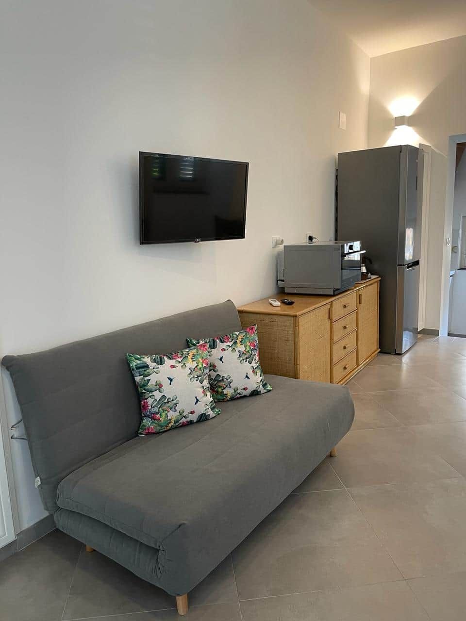 A modern living room with a grey sofa, a flat-screen TV mounted on the wall, a wooden cabinet, and a refrigerator.