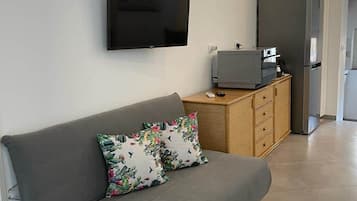 A modern living room with a grey sofa, a flat-screen TV mounted on the wall, a wooden cabinet, and a refrigerator.