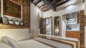 Un dormitorio con cama, mesita de noche, ventana con vistas, una zona de cocina y una pared de piedra.