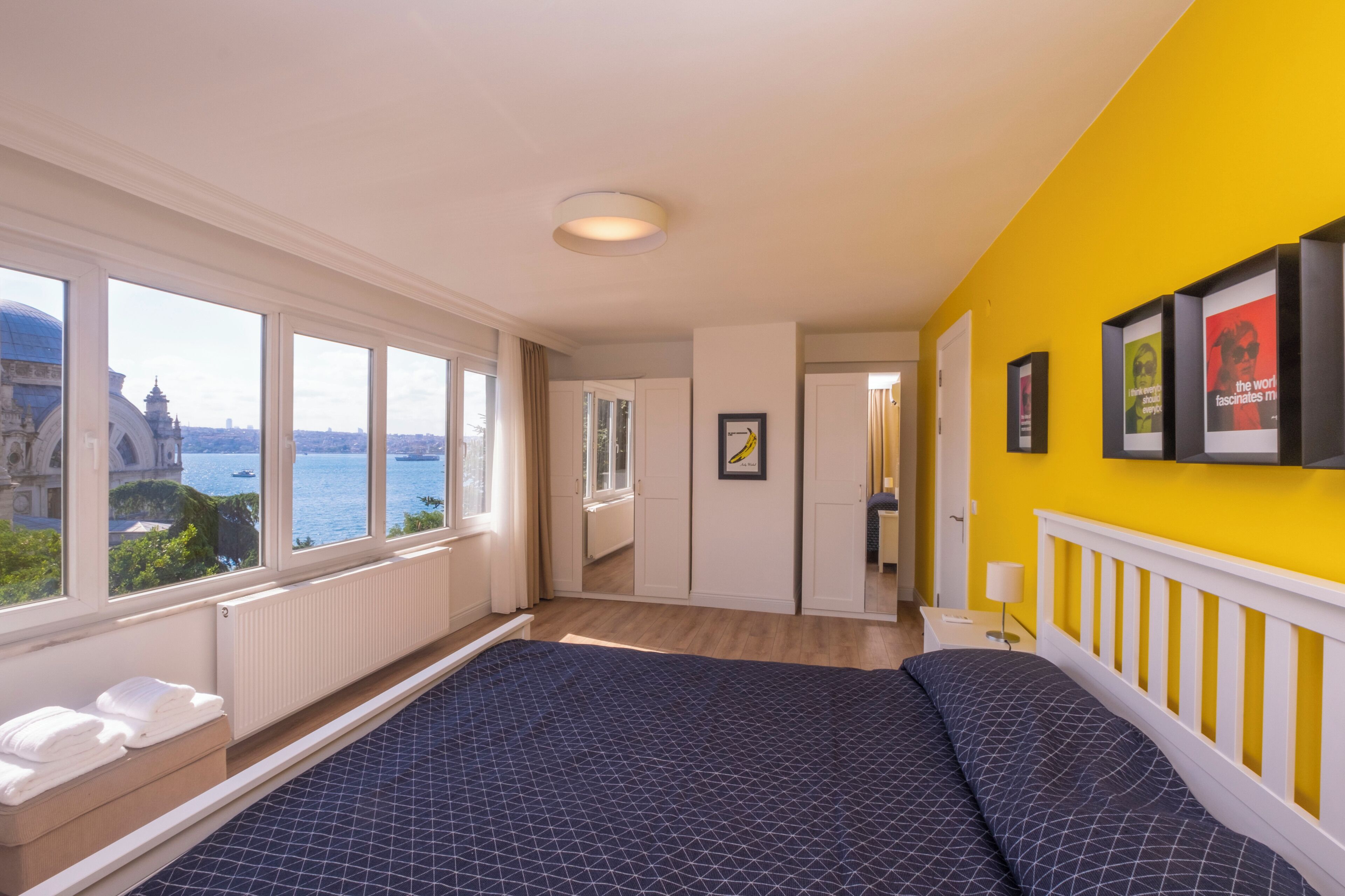 A hotel room with a large bed, a view of the sea, and a bright yellow wall.