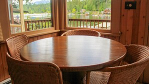 Una mesa redonda de madera con sillas de mimbre en una habitación con paredes de madera y ventanas amplias con vistas a un lago y un bosque.