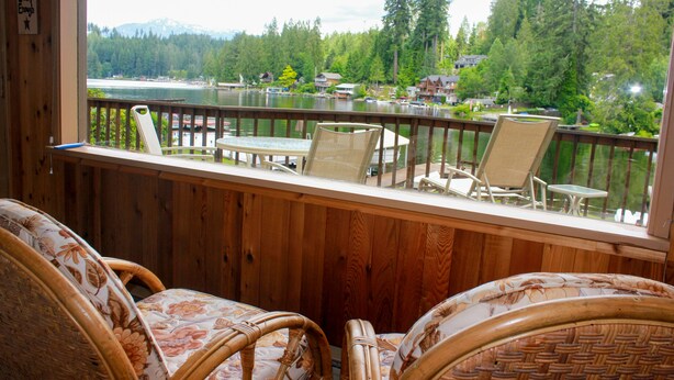 Una terraza de madera con sillas de mimbre y una mesa, con vistas a un lago y casas y árboles al fondo.