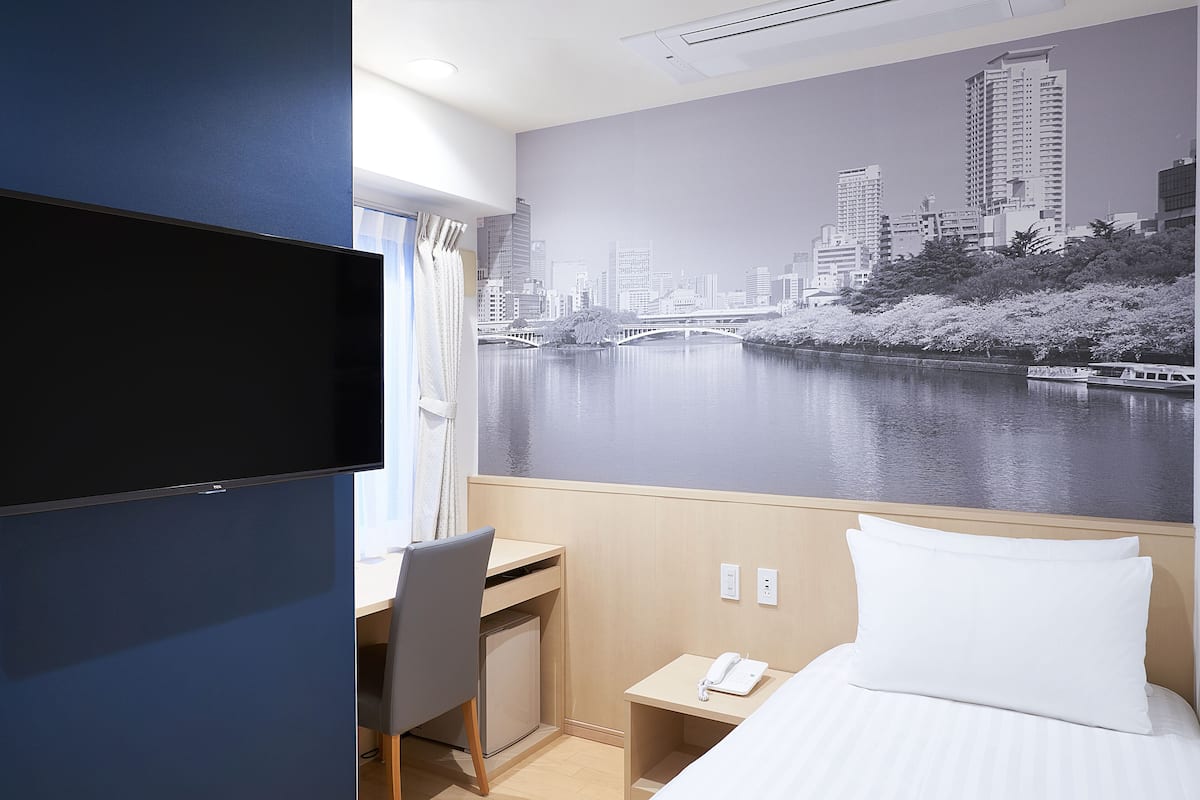 Habitación de hotel con cama, escritorio, silla, mesita de noche pequeña con teléfono y un mural en la pared que muestra un paisaje urbano con un río.
