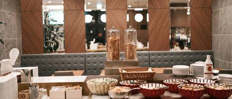 A buffet setup with various food items and serving dishes, a beverage dispenser, and a seating area in the background.