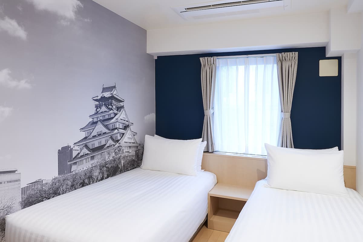 Habitación de hotel con dos camas, un ventanal con cortinas, un mural en la pared de una construcción tradicional y un respiradero en el techo.