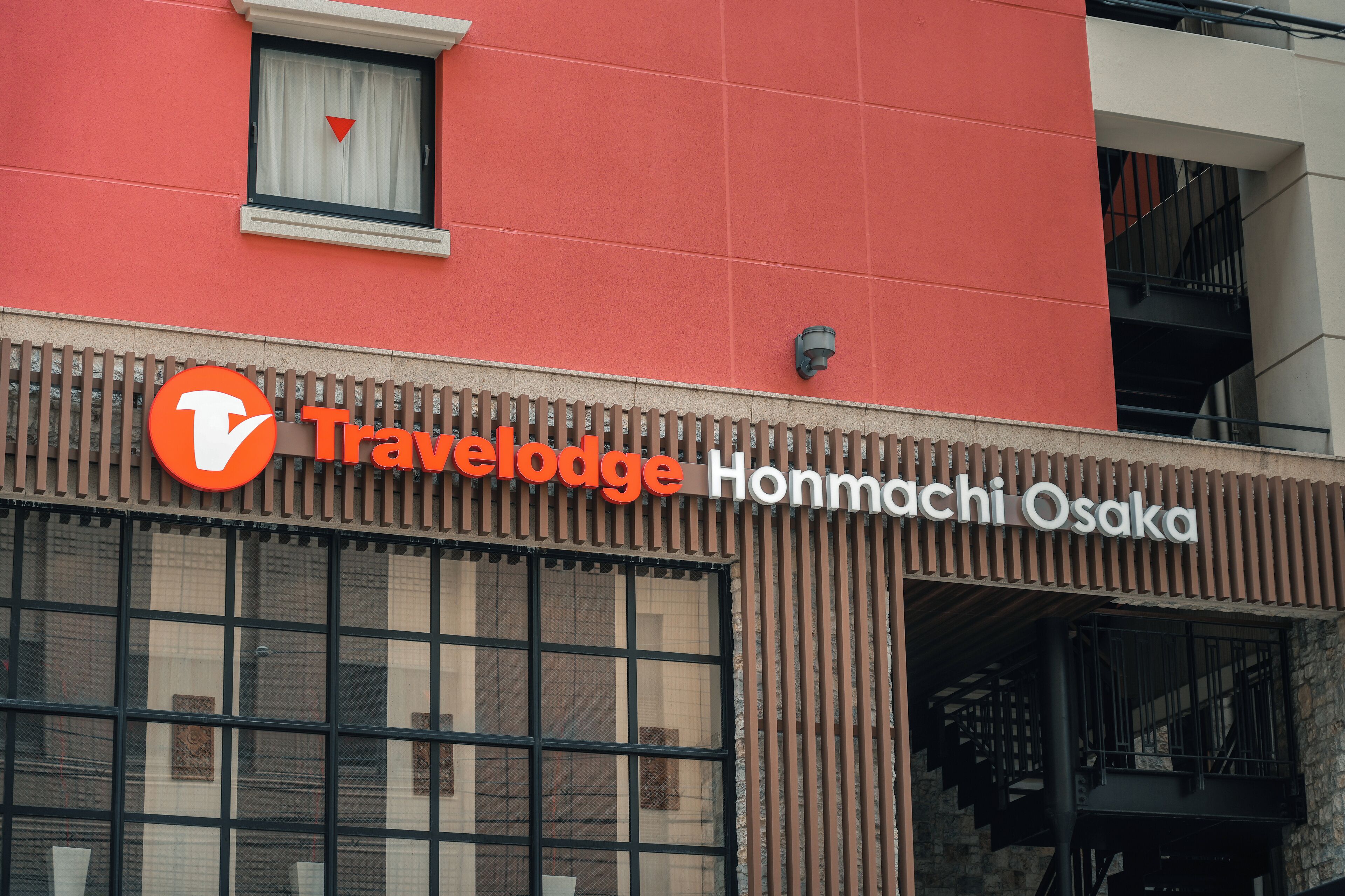 Un letrero de Travelodge con un logotipo rojo y blanco, ubicado sobre la entrada de un edificio con fachada marrón y una ventana.