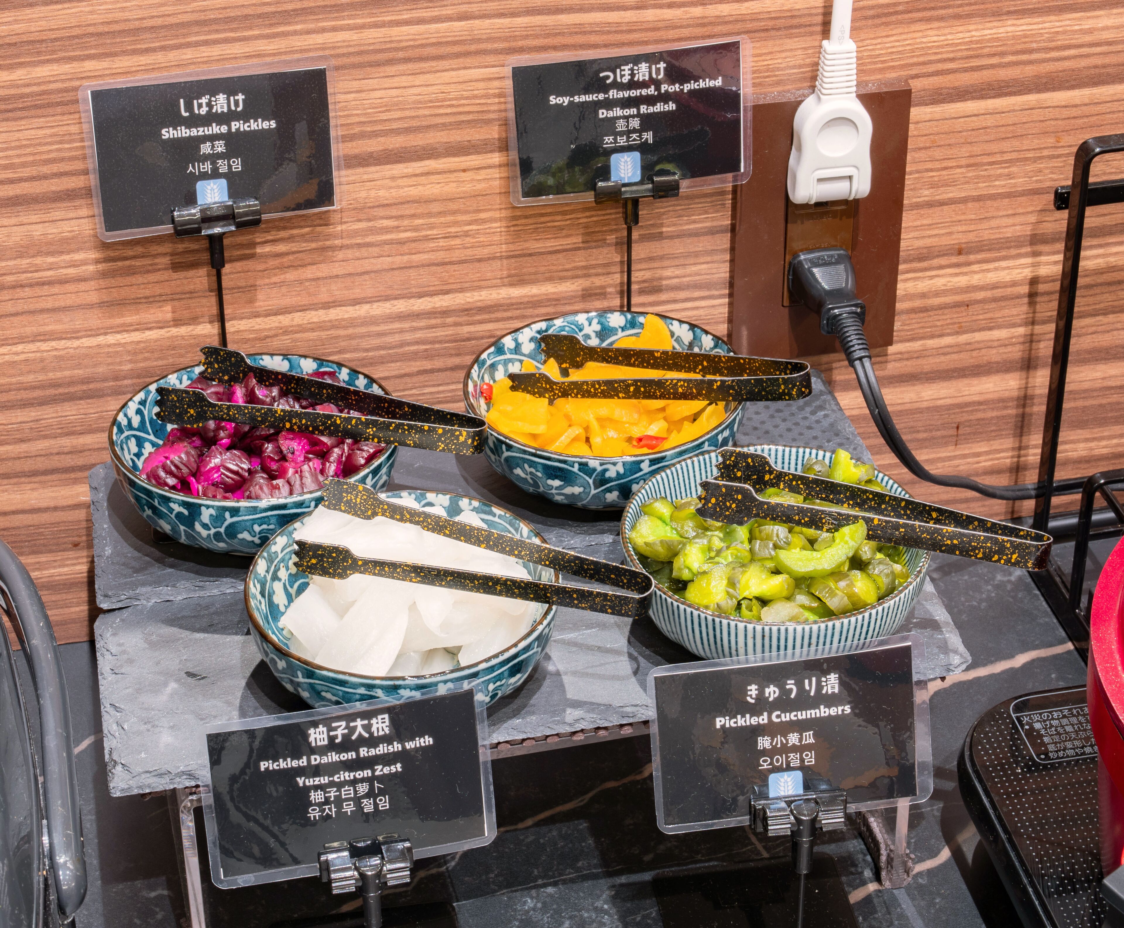 A food display with various pickled items, including Shibazuke Pickles, Daikon Radish, and Pickled Cucumbers, each labeled with their names and descriptions.