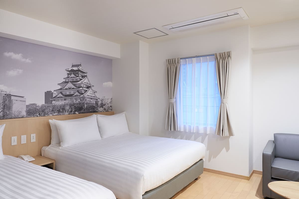 Habitación de hotel con cama, una silla, una mesita, ventana con cortinas y un mural en la pared con un castillo.