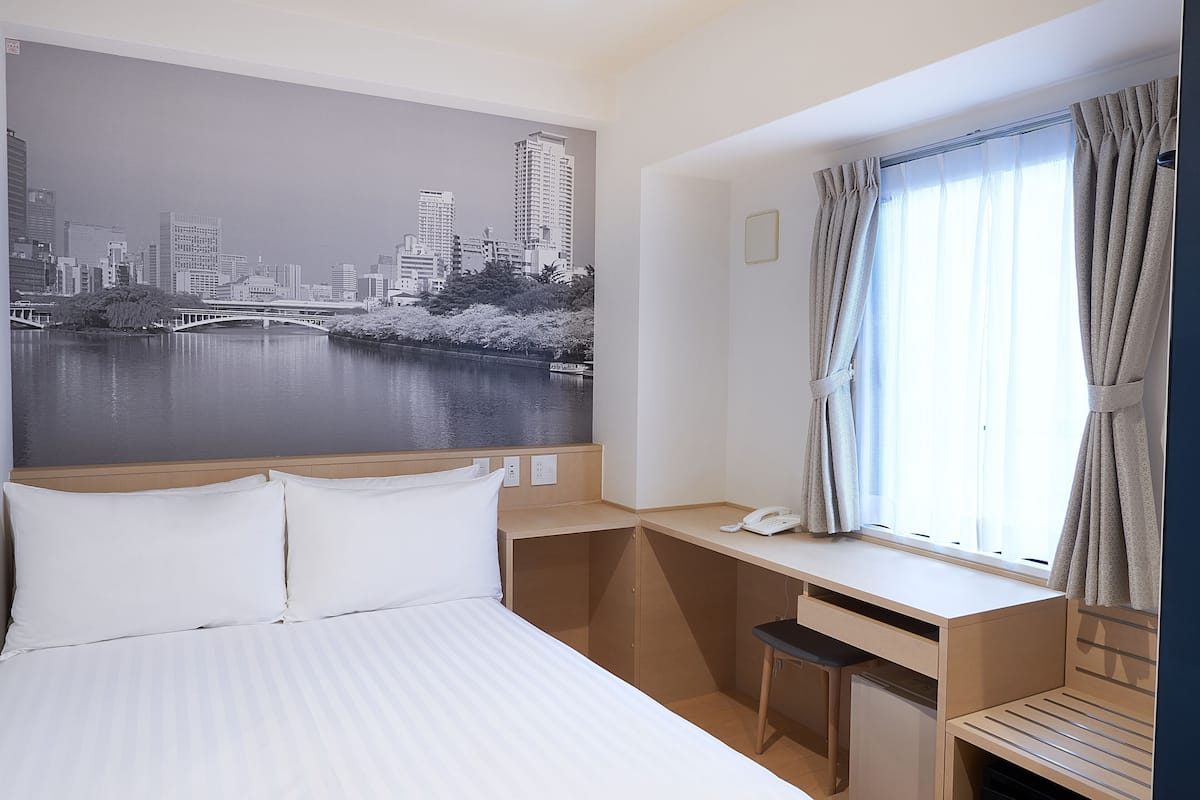Habitación de hotel con cama, escritorio, silla, ventana con cortinas y un cuadro de paisaje urbano en la pared.