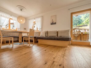 A modern living room with a wooden floor, a sofa, a dining table with chairs, and a large window with a view of a mountainous landscape.