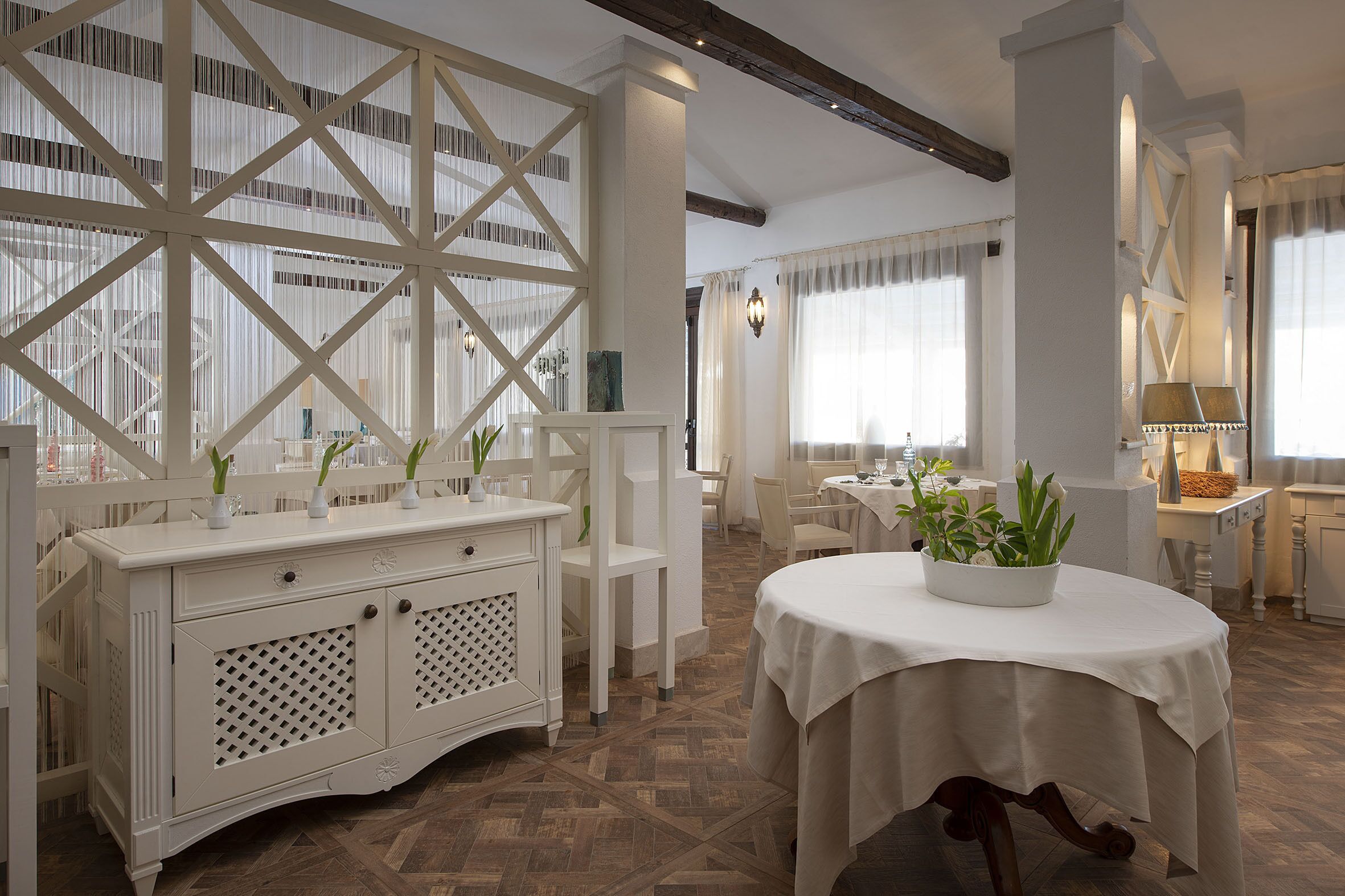 Una habitación de hotel espaciosa con un ventanal grande, una consola blanca con plantas decorativas, una mesa de comedor redonda y suelo de madera.