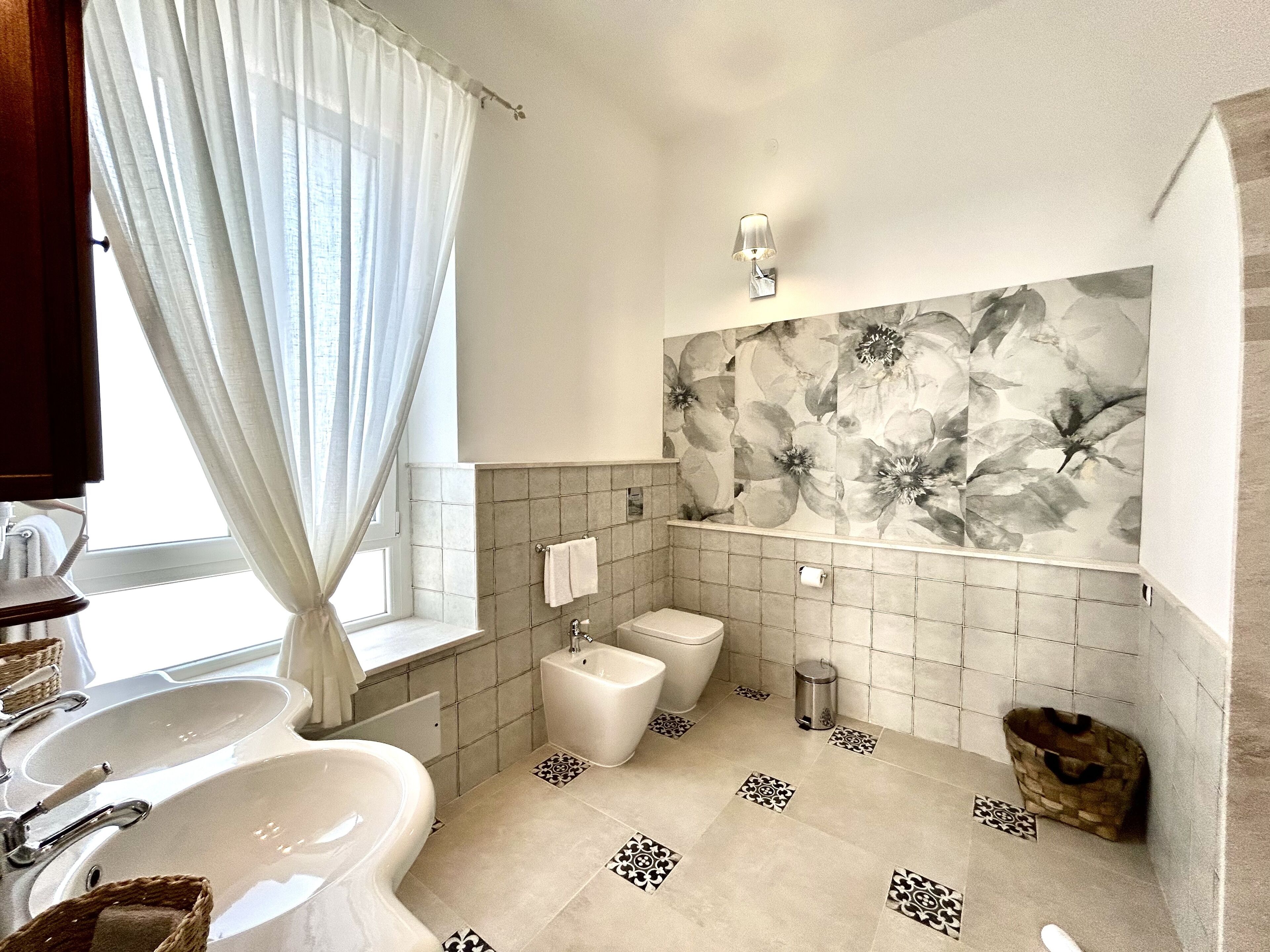Un baño moderno con un lavamanos grande, un bidé y un inodoro. Hay una pared decorativa con un estampado floral y una ventana con cortinas traslúcidas.
