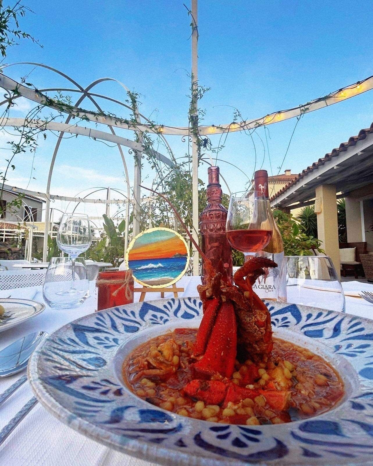 Un plato de mariscos sobre una mesa, con vista a un atardecer pintado, una botella de vino tinto y un vaso de agua.