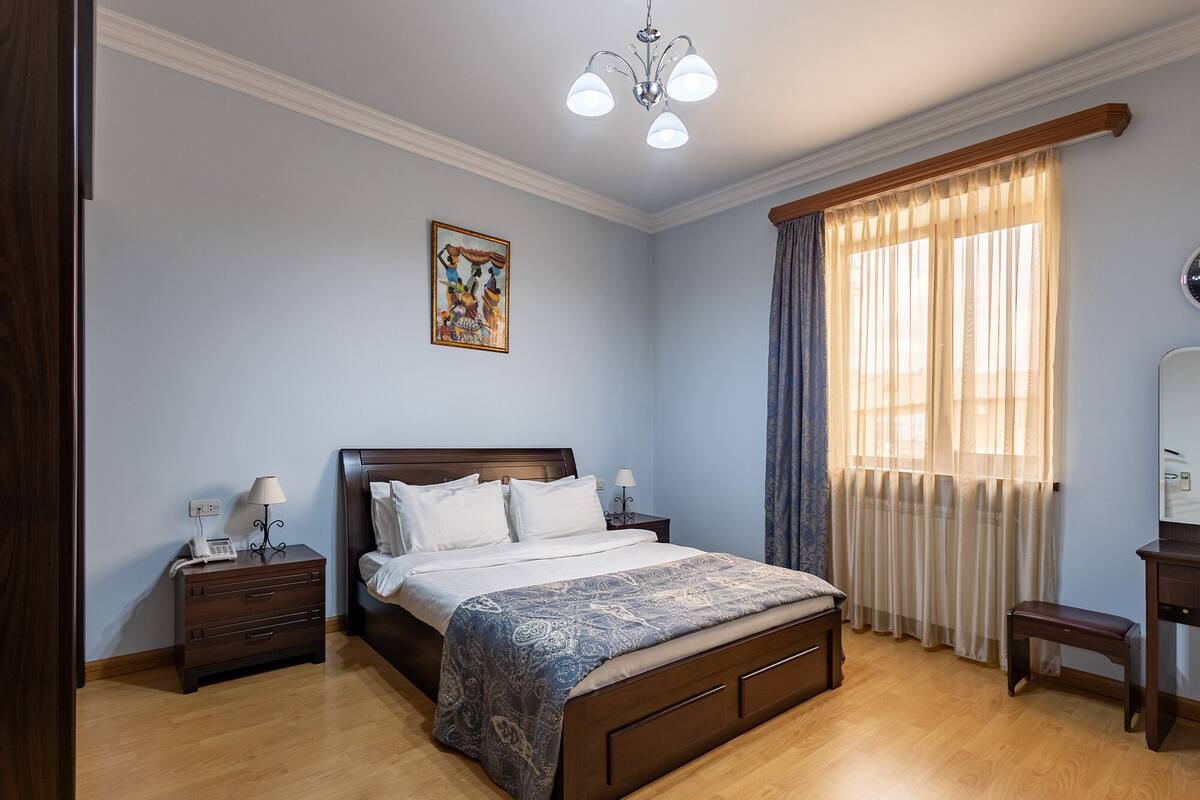 A bedroom with a wooden bed, bedside tables, a nightstand, a chair, a window with curtains, and a ceiling light fixture.