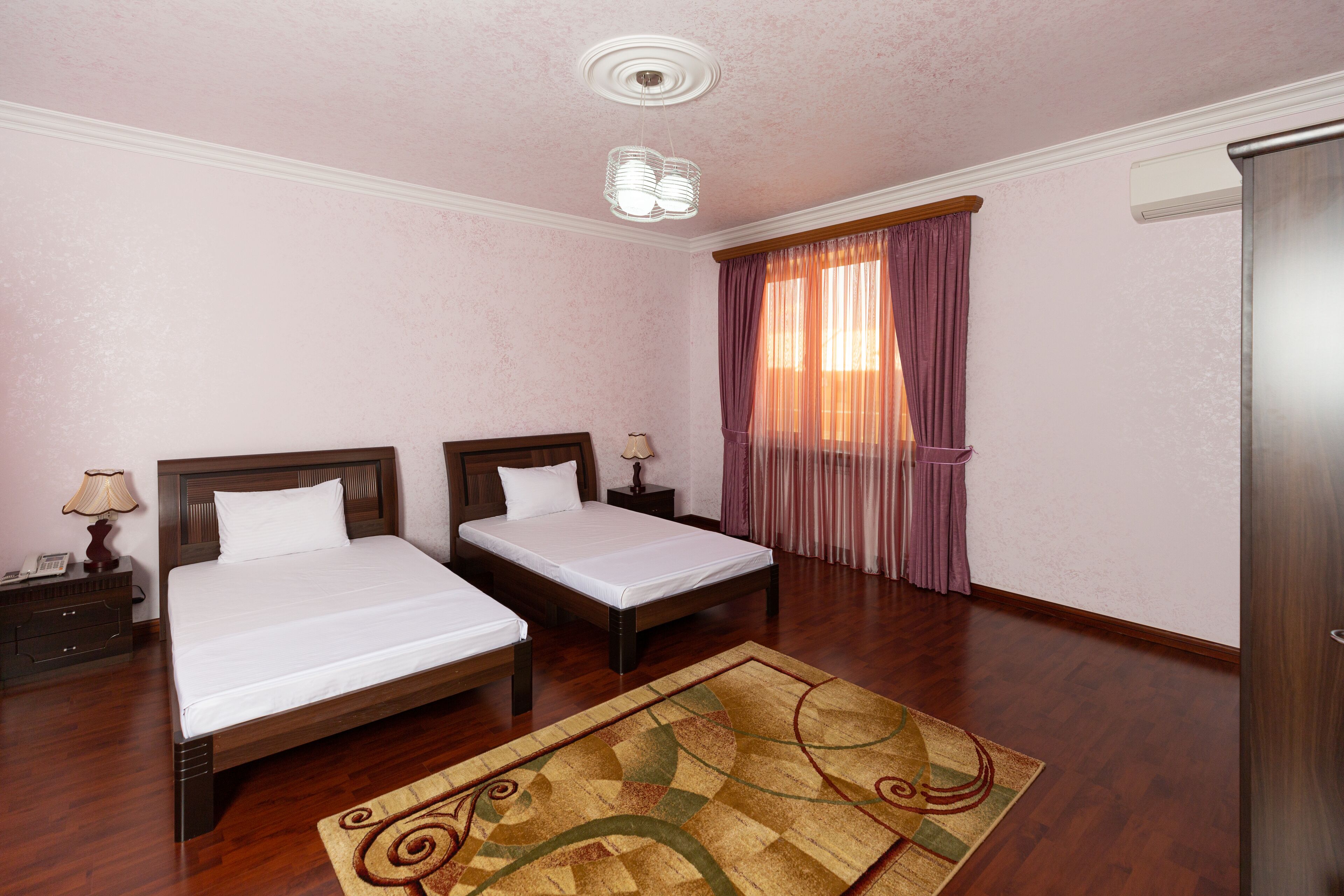 A hotel room with two beds, a wooden floor, a patterned rug, a ceiling light fixture, and a window with curtains.