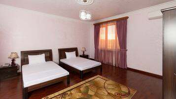 A hotel room with two beds, a wooden floor, a patterned rug, a ceiling light fixture, and a window with curtains.