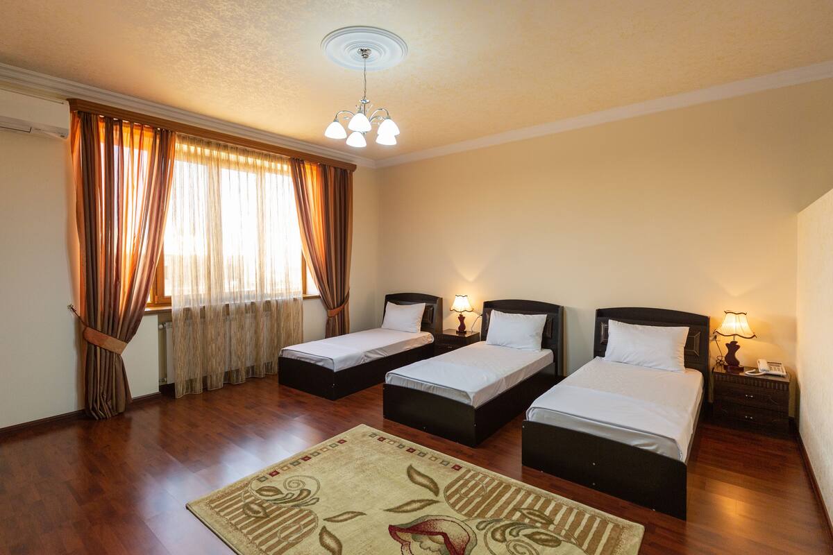 A hotel room with three single beds, wooden flooring, a large window with curtains, a ceiling light fixture, and a small rug.
