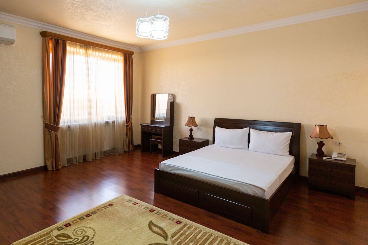 A hotel room with a large bed, wooden floor, a desk with a chair, a nightstand with a lamp, and a window with curtains.