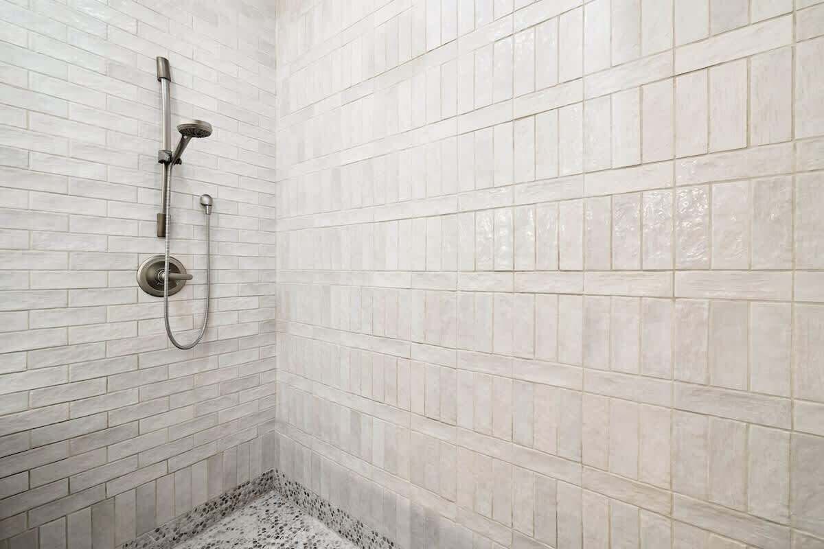 A shower with a handheld showerhead and a wall-mounted faucet, tiled with light-colored tiles.