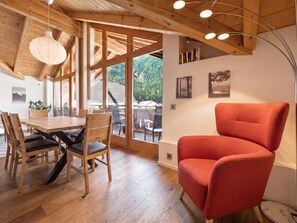 A dining area with a wooden table and chairs, a red armchair, and a view through large windows.