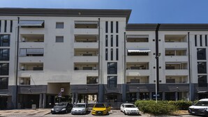A multi-story residential building with balconies, a parking area, and a clear sky.