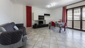 A modern living room with a grey sofa, a dining table with red chairs, a TV, and a balcony with red curtains.