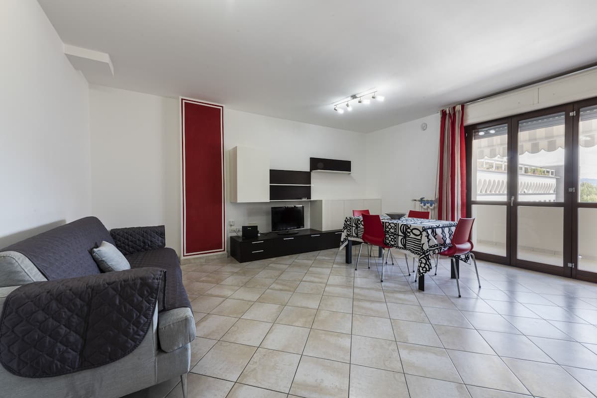 Una sala de estar moderna con un sofá gris, una mesa de comedor con sillas rojas, un televisor y un balcón con cortinas rojas.
