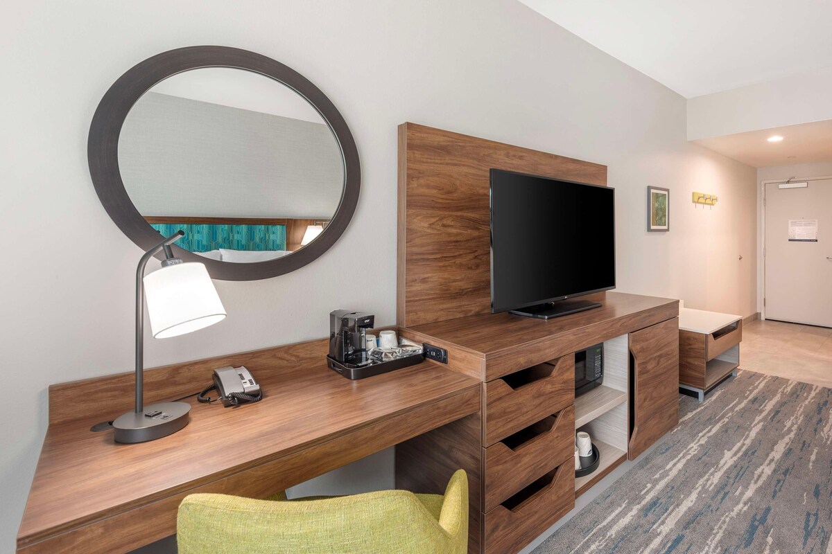 Ein Hotelzimmer mit Schreibtisch aus Holz, einem Flachbildfernseher, einem Spiegel, einer Lampe, einem Telefon und einem grünen Sessel.