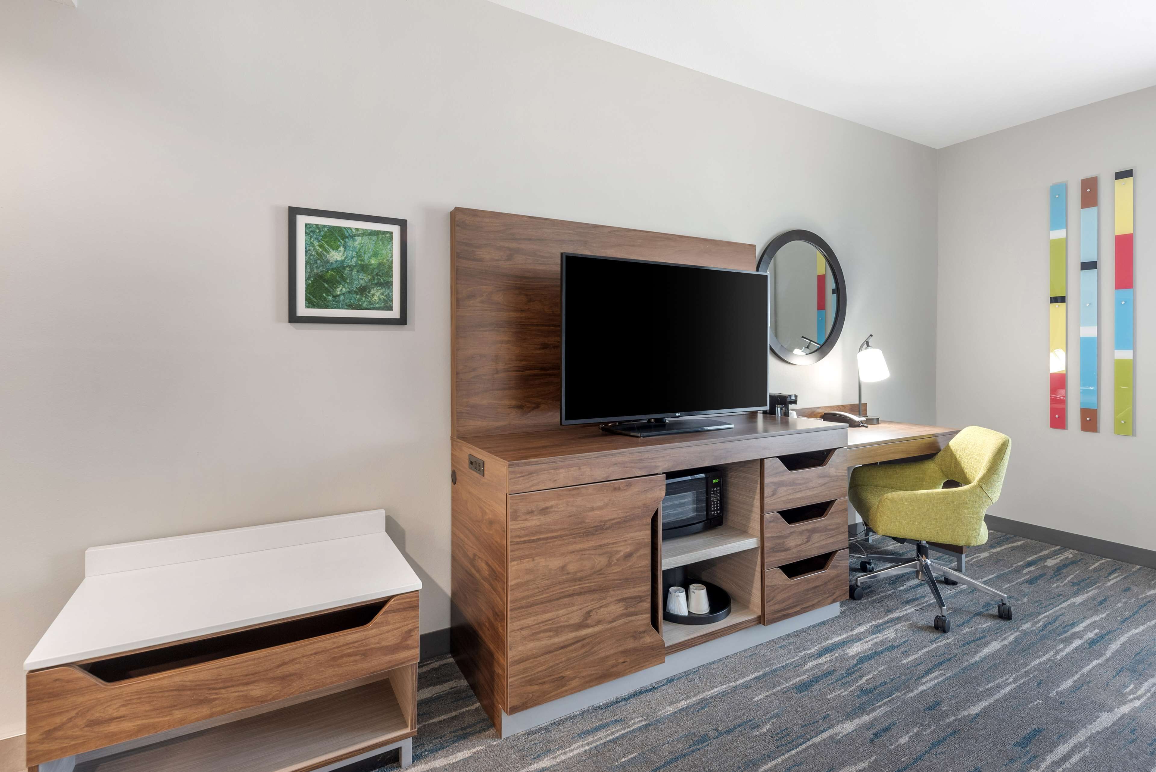 A hotel room with a wooden desk, a flat-screen TV, a mirror, a yellow chair, and a framed picture on the wall.
