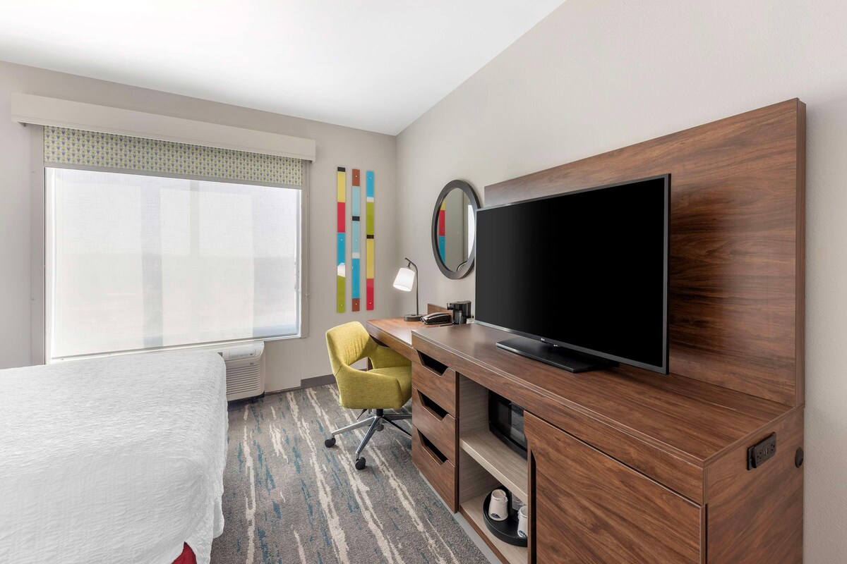 A hotel room with a bed, a desk with a television, a chair, a mirror, and a window with blinds.