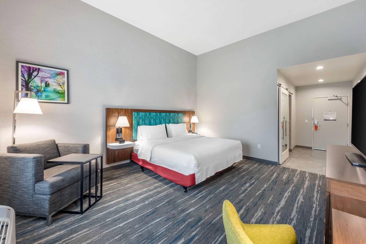 A hotel room with a bed, two bedside tables with lamps, a sofa, a chair, a TV, and a painting on the wall.