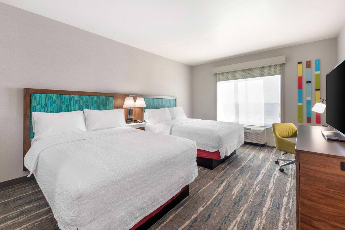 A hotel room with two beds, a desk, a chair, and a large window.